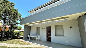 A modern two-story building with a flat roof, a covered patio with a table and chairs, and a door leading to the interior. - Bright Flat With Shared Pool (San Michele al Tagliamento)