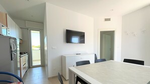 A modern kitchen with a refrigerator, stove, and cabinets. There is a dining area with a table and chairs. A television is mounted on the wall. - Great and Bright Flat With Pool (San Michele al Tagliamento)