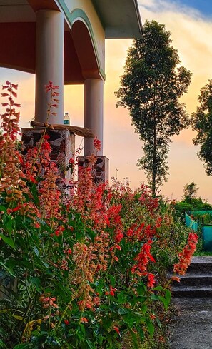 A covered outdoor area with a column structure, surrounded by flowering plants and a tree. - Infinity Viewpoint Homestay (Kalimpong)