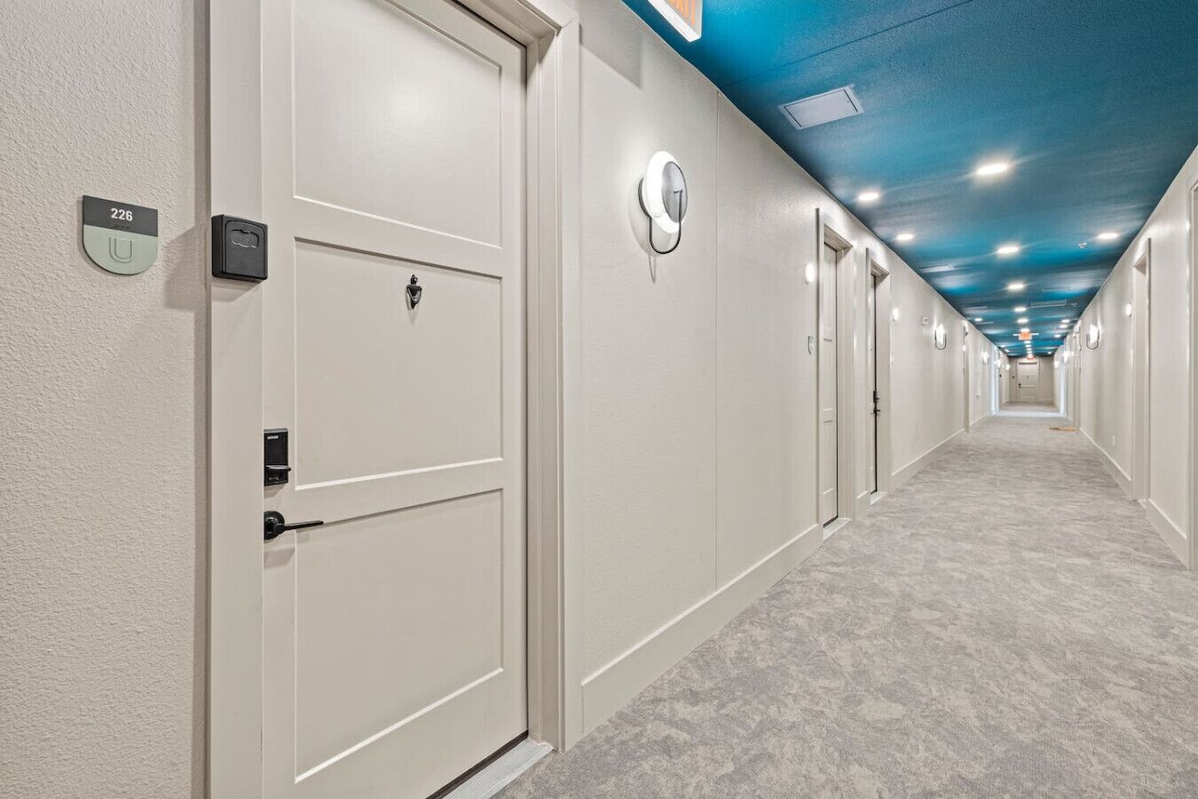 A hotel hallway with white doors, numbered room signs, and a blue ceiling.