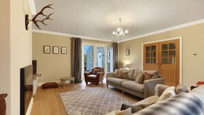 Ein geräumiges Wohnzimmer mit einer Couch, Sesseln, einem Couchtisch und einer Chandelüster. Eine Glastür bietet Blick ins Freie.
