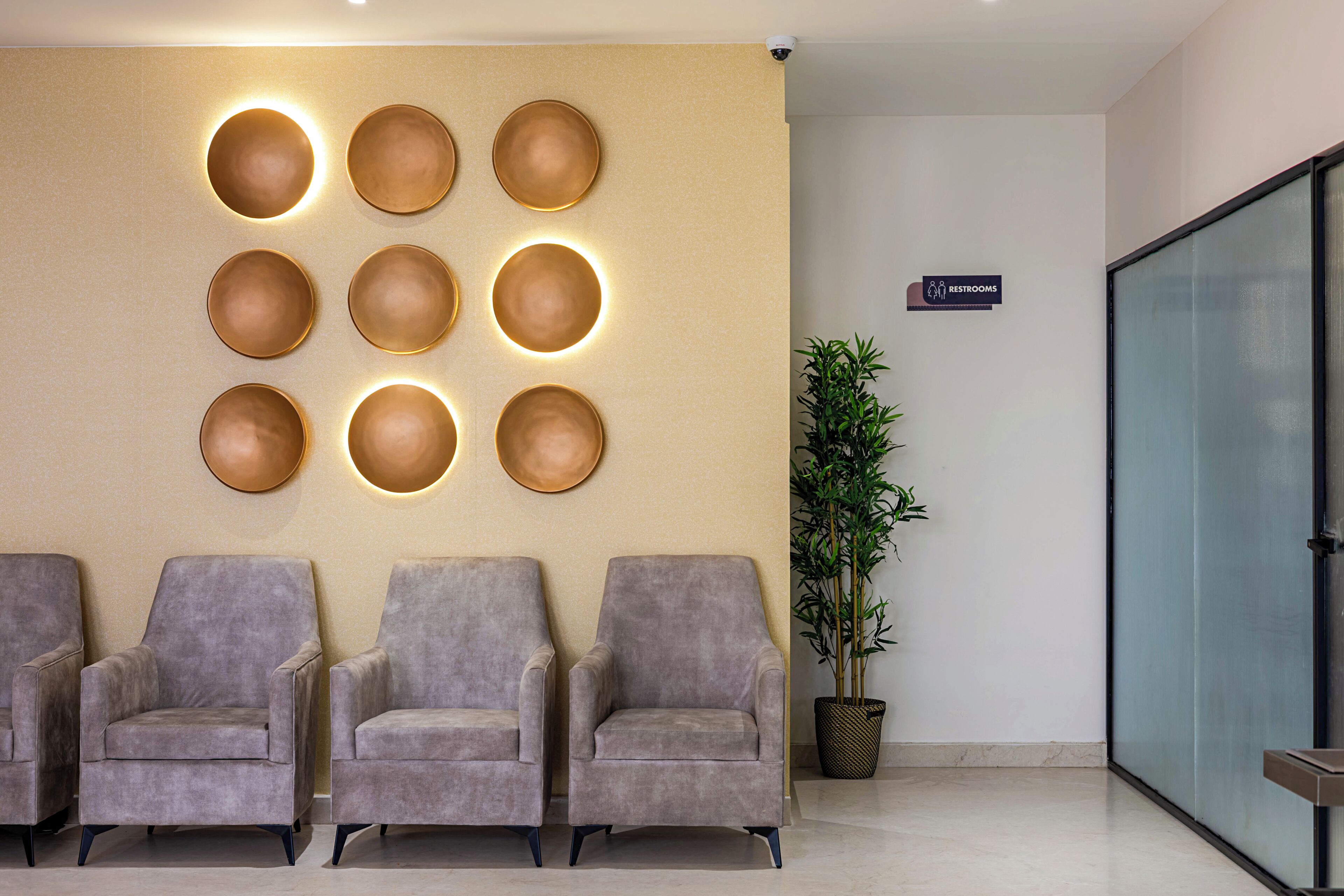 Un espace d’accueil avec un luminaire fixé au mur, un panneau indiquant l’emplacement des toilettes, trois fauteuils gris et une plante en pot.