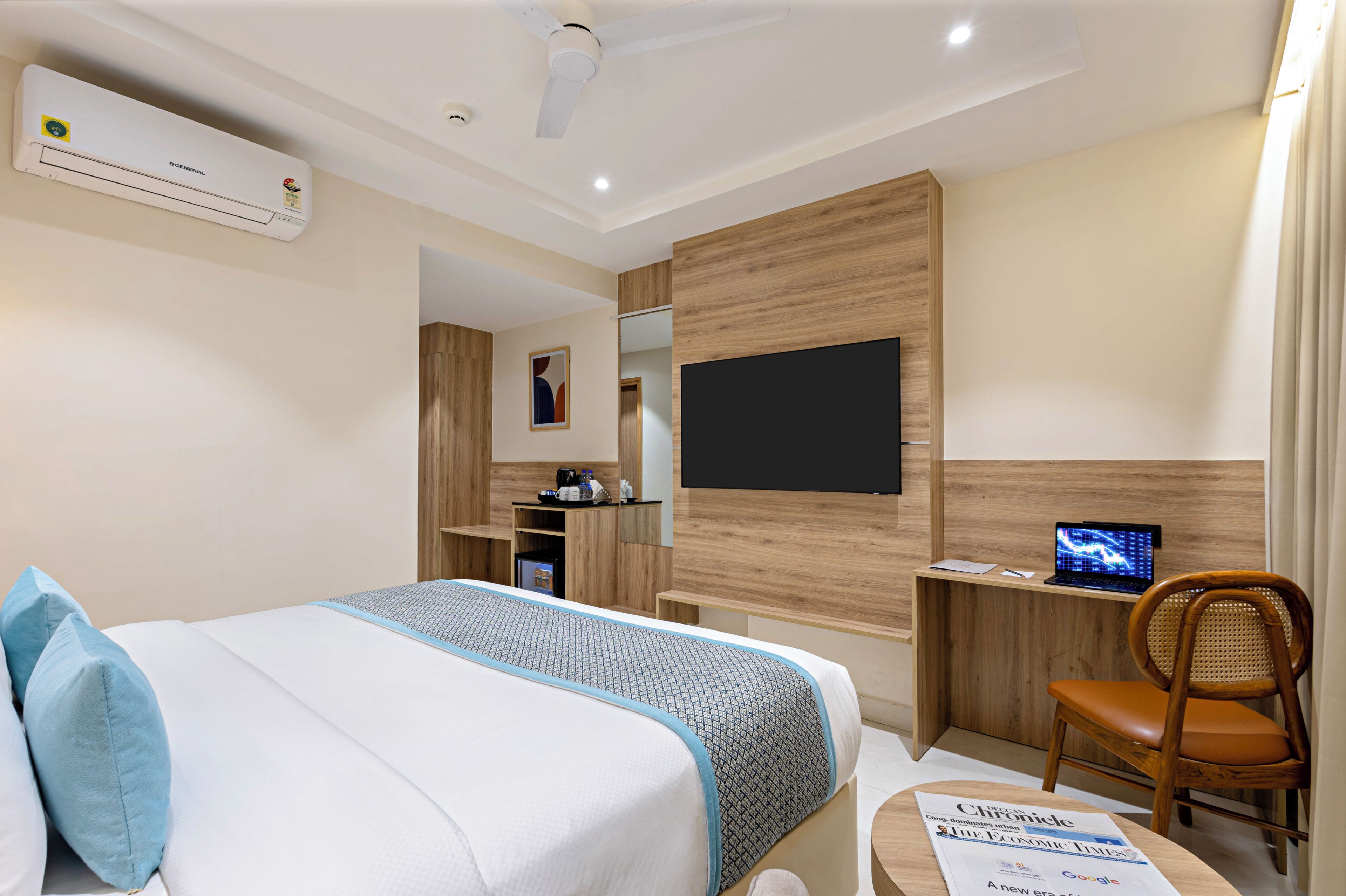 A hotel room with a bed, a desk with a computer, a chair, a flat-screen TV, and an air conditioning unit.