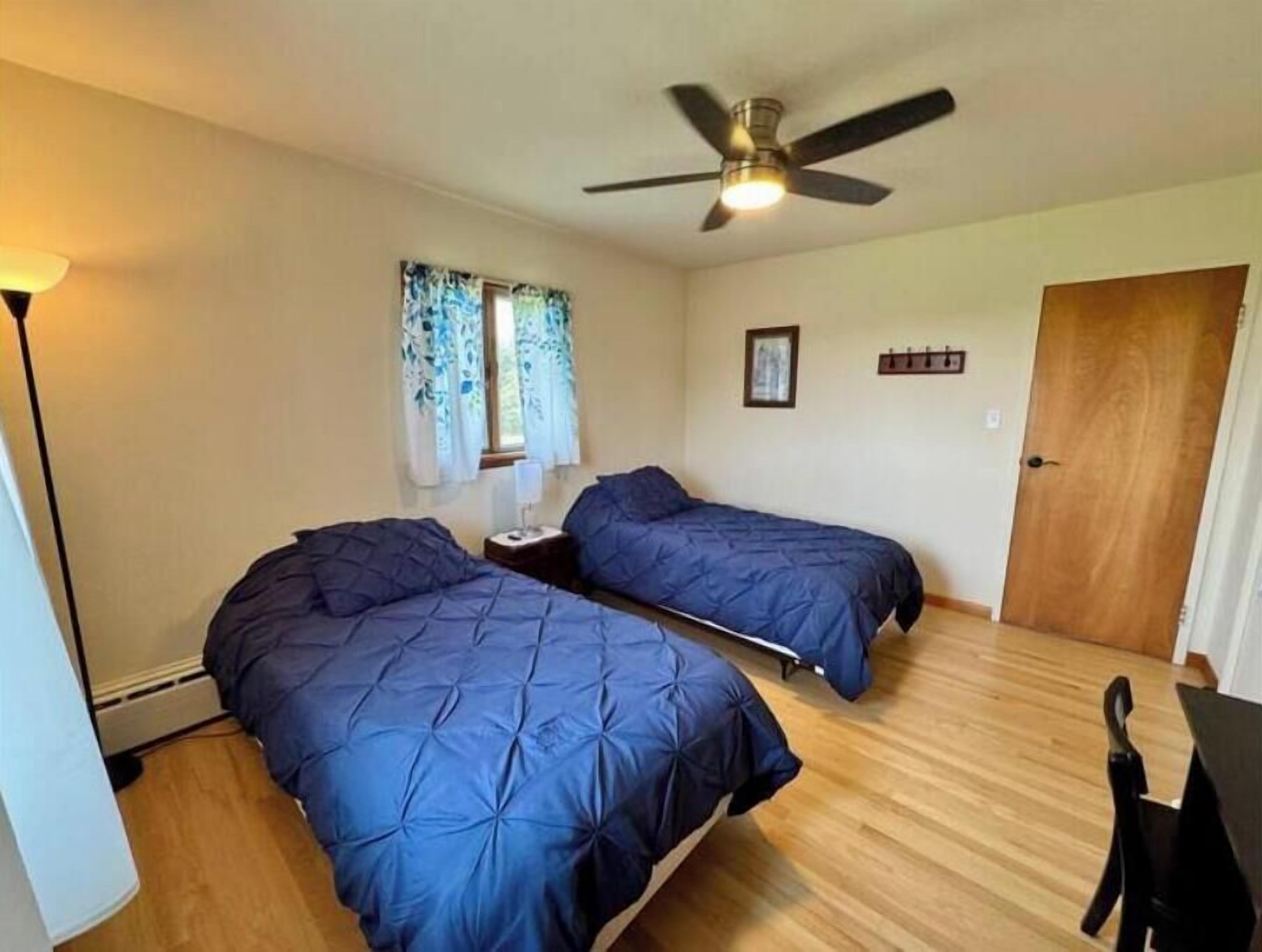 Une chambre avec deux lits, un ventilateur de plafond, une fenêtre avec des rideaux, une porte et une petite table.