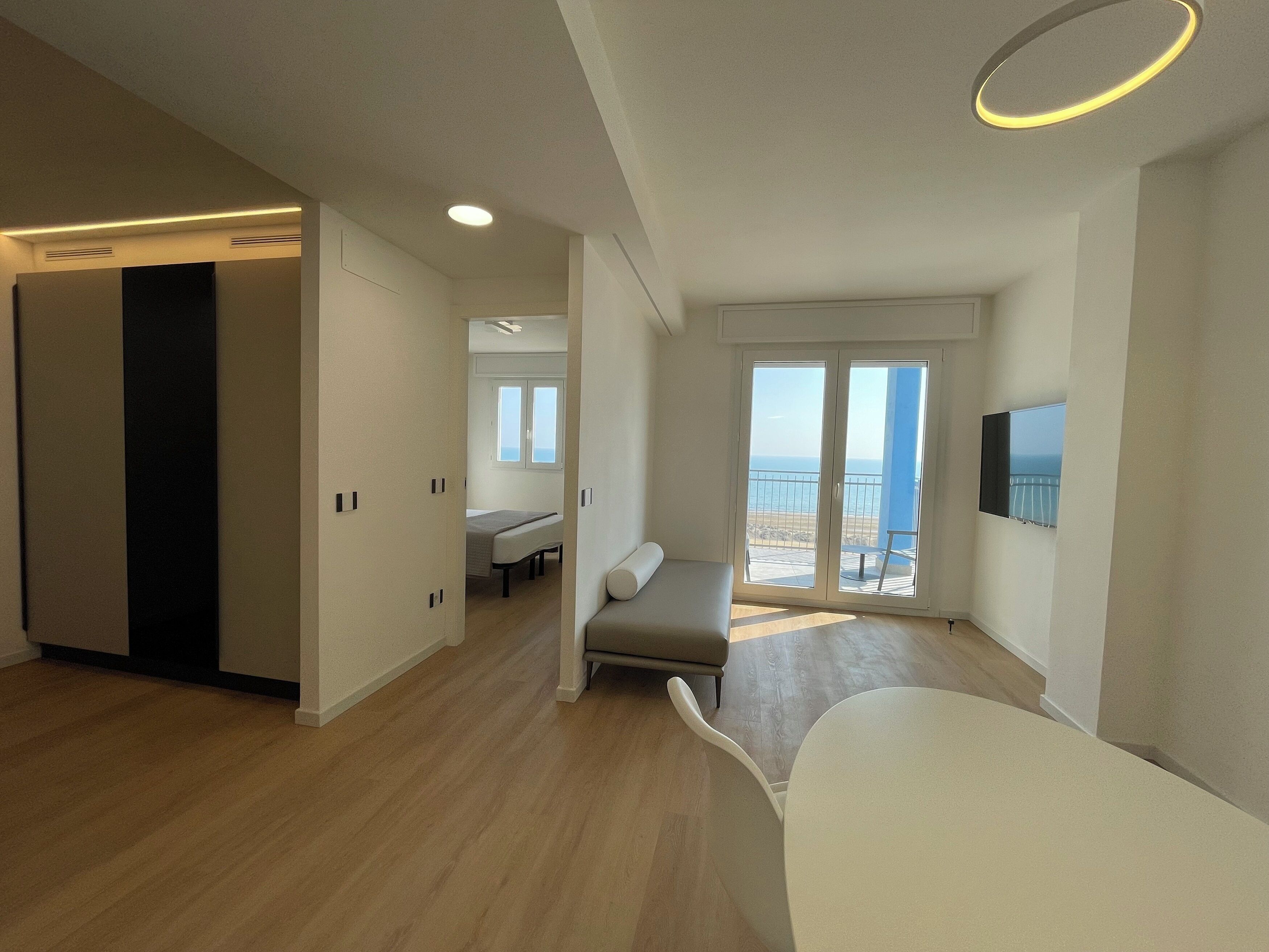 Une pièce moderne et bien éclairée, dotée d’une grande fenêtre donnant sur la mer, d’un téléviseur à écran plat fixé au mur et d’un coin salon avec un canapé et une petite table.