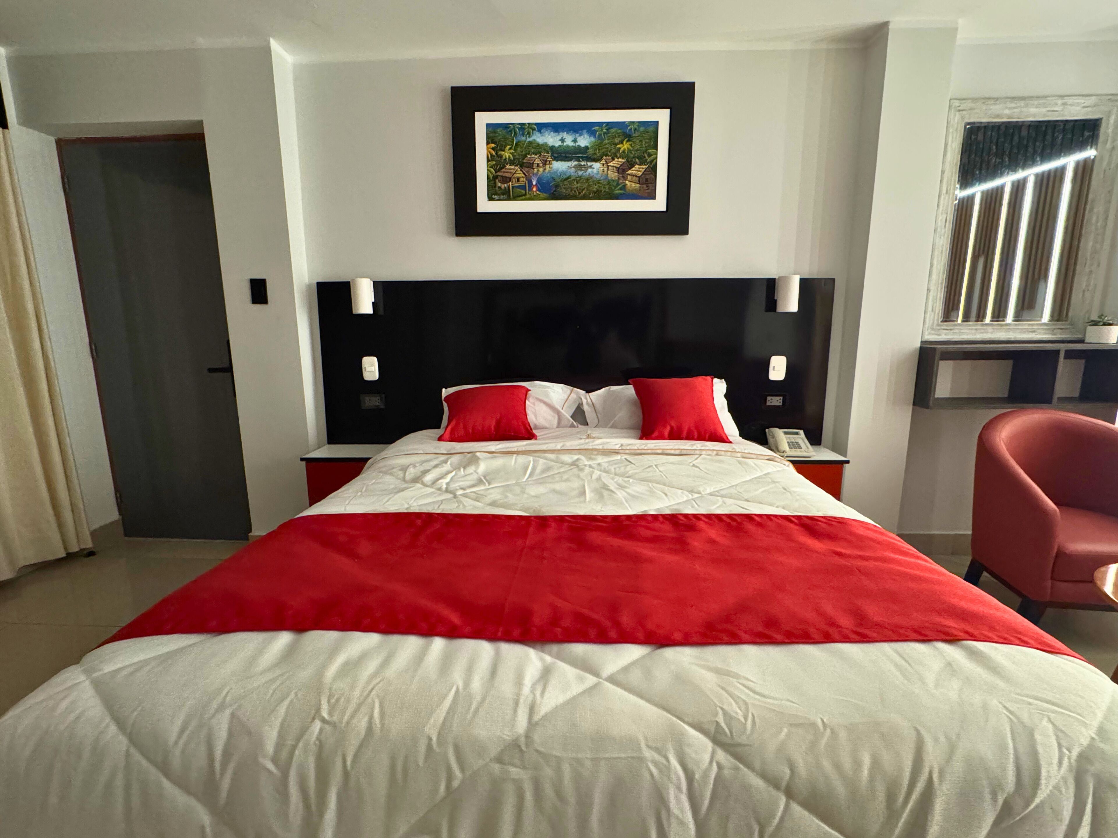 Habitación de hotel con una cama grande, un cuadro enmarcado en la pared, una mesita de noche con un teléfono y una silla roja.