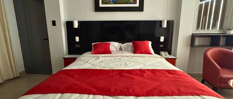 Habitación de hotel con una cama grande, un cuadro enmarcado en la pared, una mesita de noche con un teléfono y una silla roja.