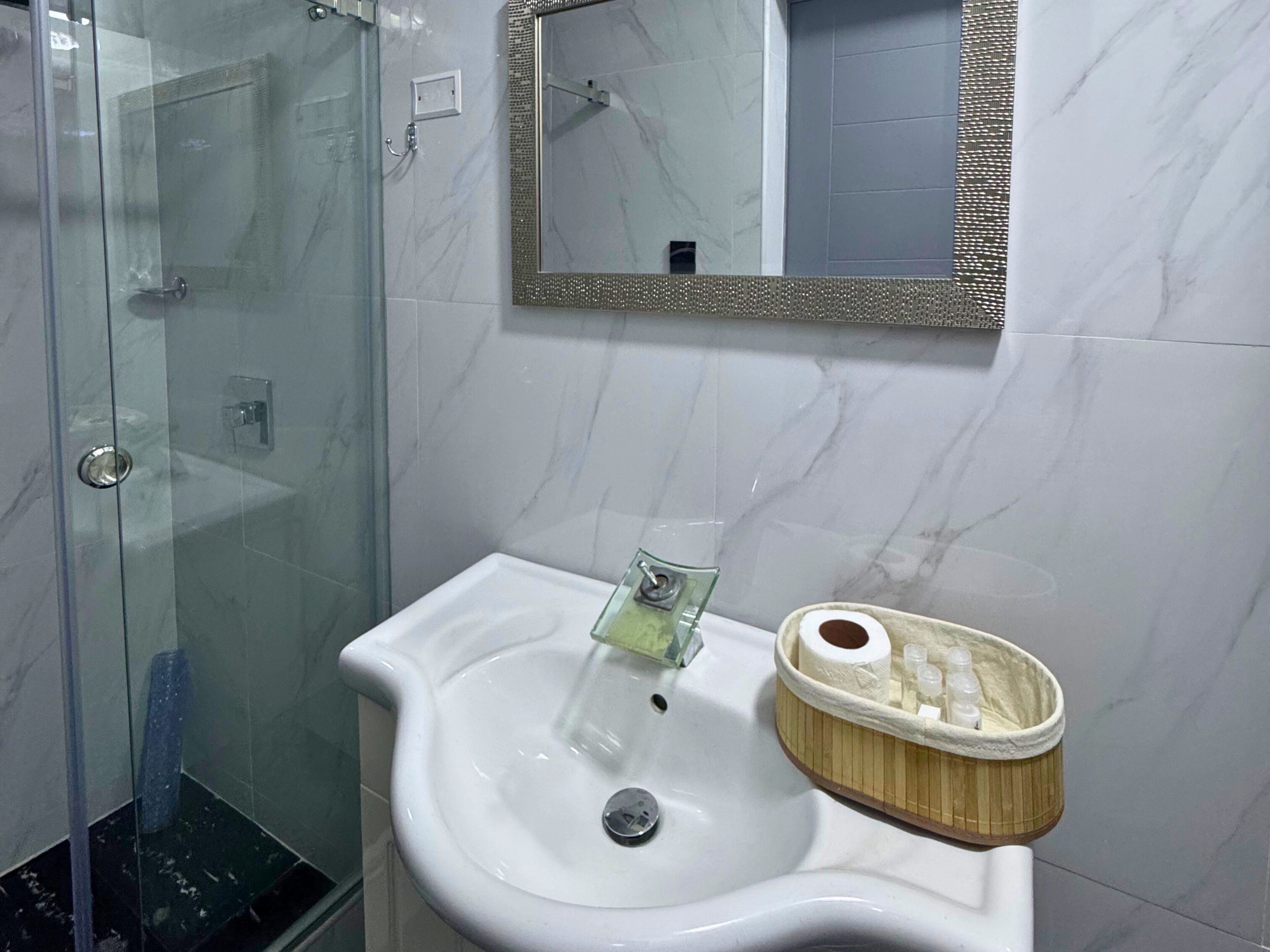 A bathroom with a glass shower enclosure, a white sink with a green faucet, and a mirror with a decorative frame.