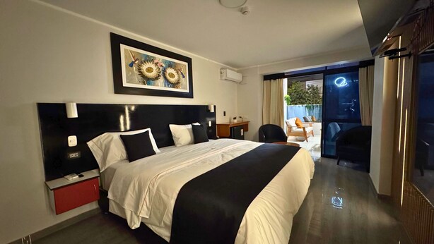 A modern hotel room with a large bed, a desk, and a chair. There is a painting on the wall and a view of the outdoors through a sliding door. - City Tower Hotel (Santiago de Surco)
