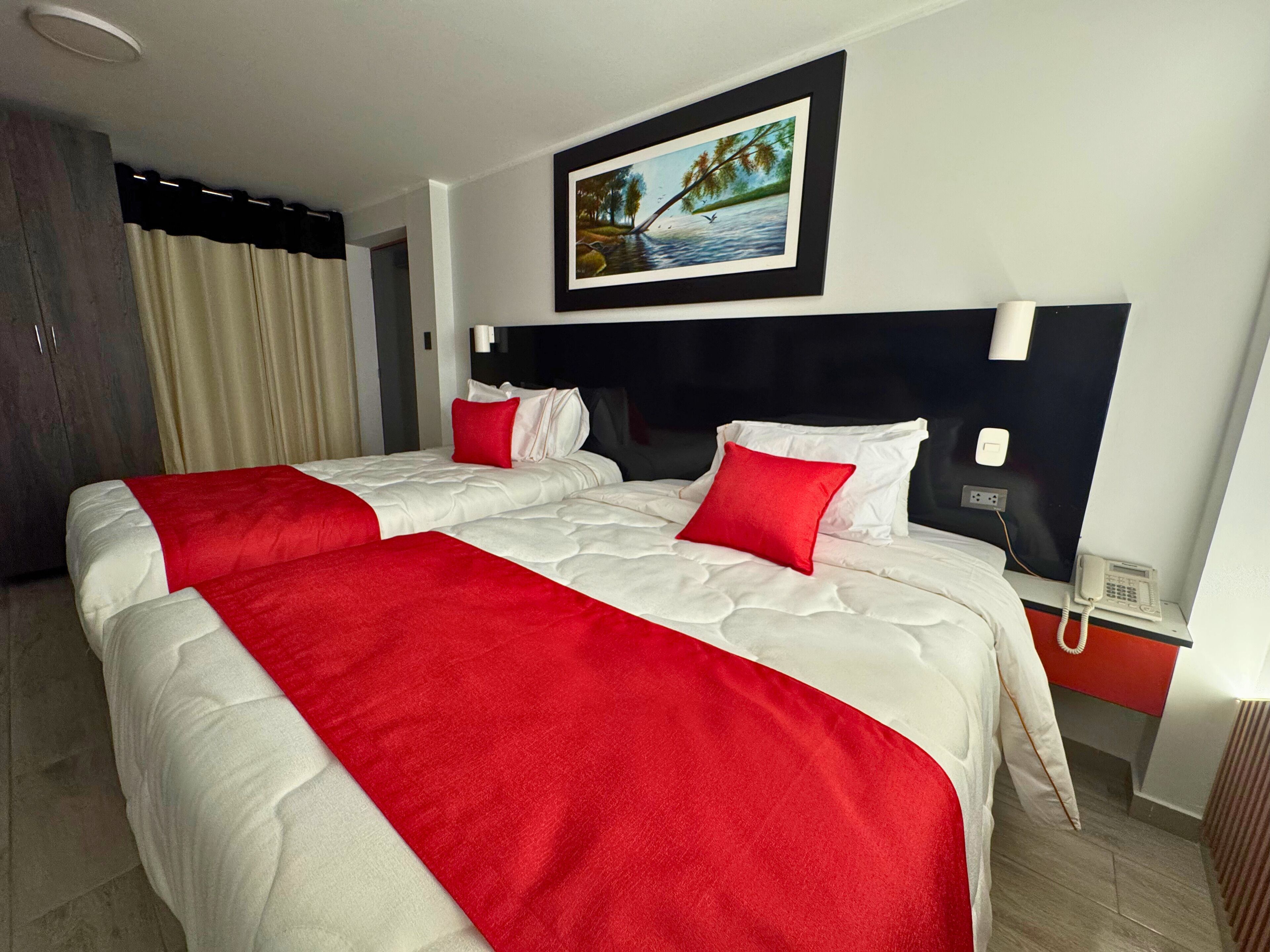 Une chambre d’hôtel avec deux lits, une literie rouge et blanche, un tableau représentant un paysage, un téléphone et une fenêtre avec des rideaux.