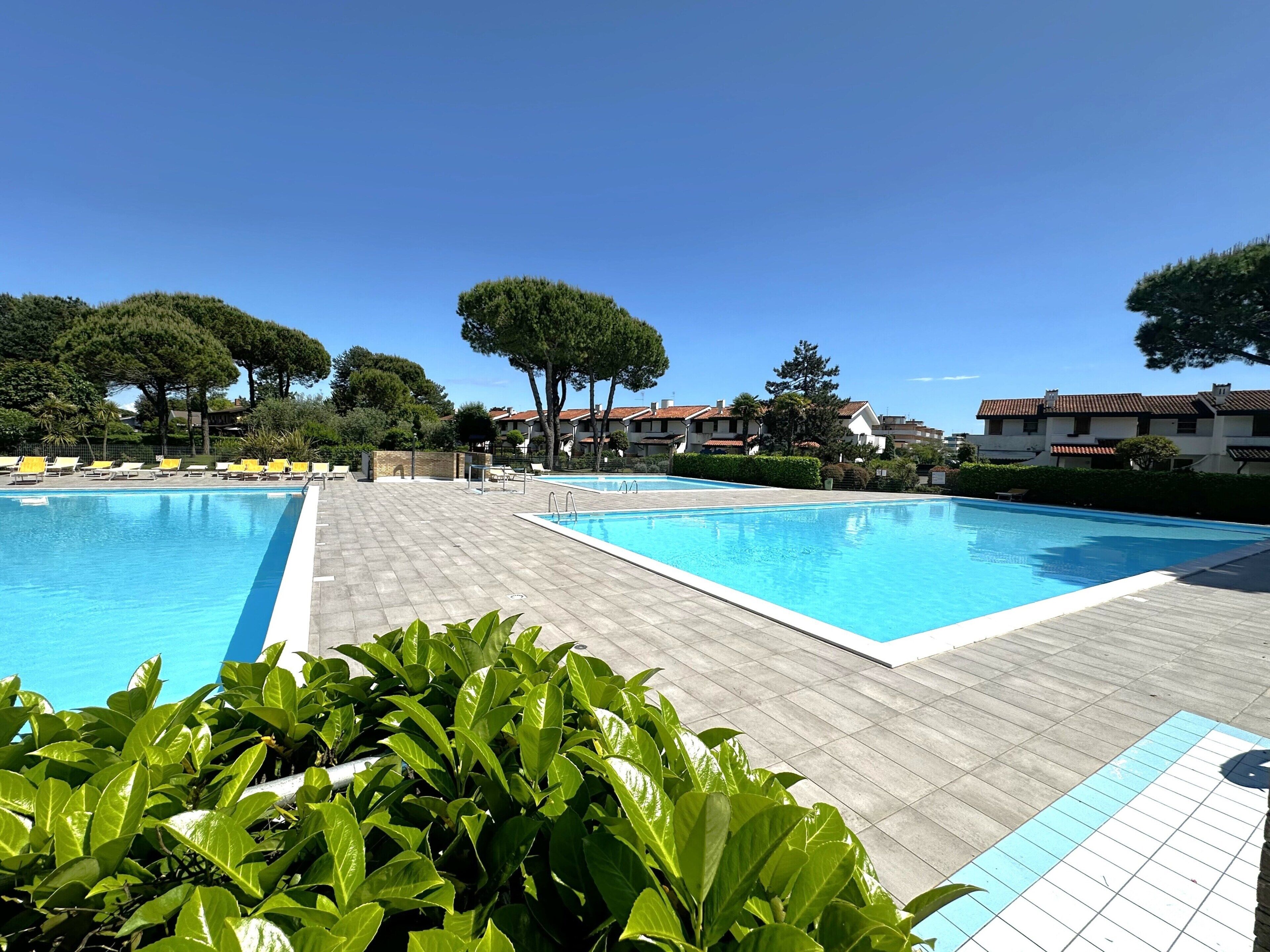 Piscina con dos zonas separadas, rodeada por un solárium de madera y sillas reclinables amarillas.