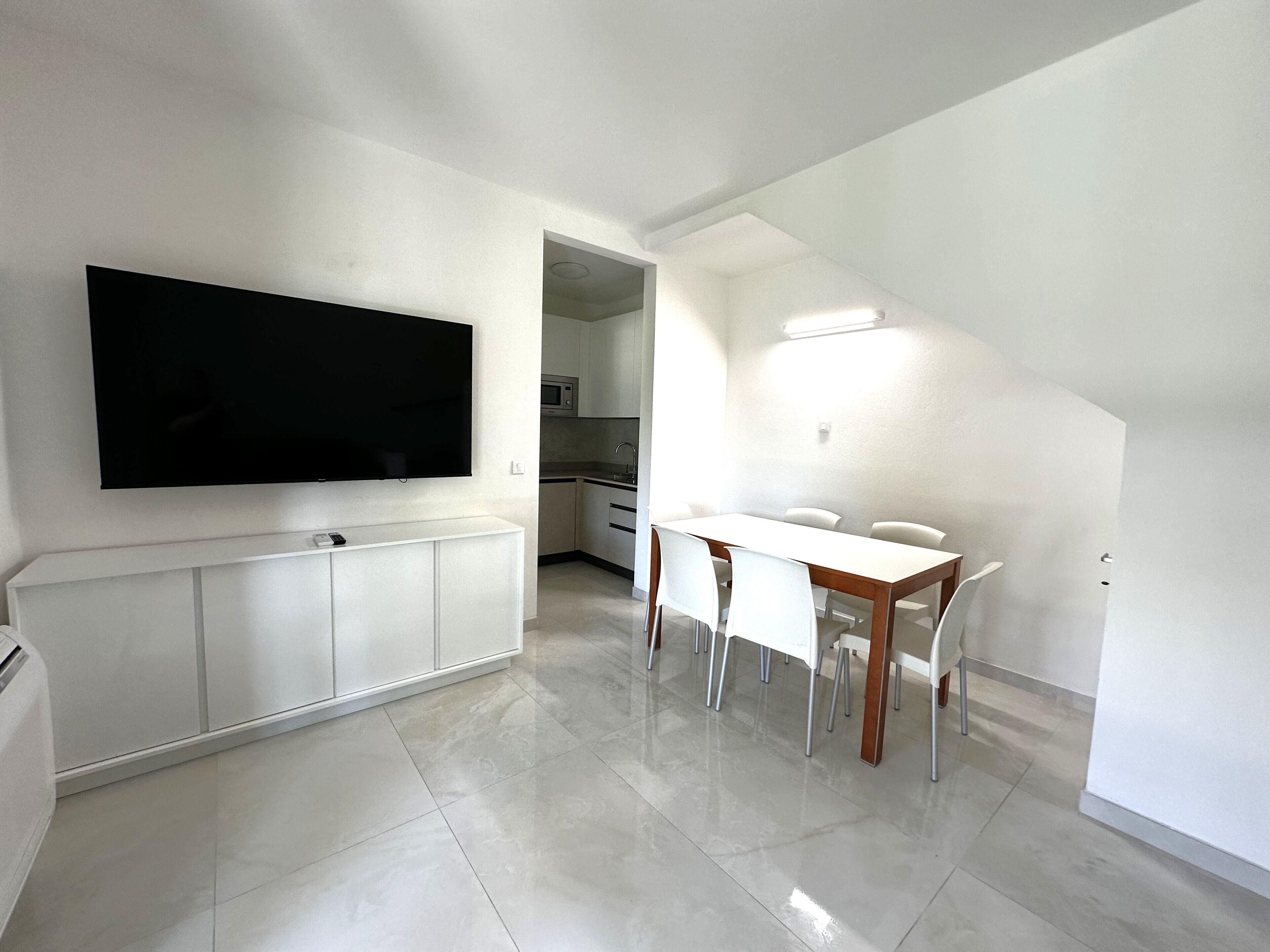 Een moderne keuken met witte kastjes, een flatscreen televisie boven de aanrecht en een eethoek met een witte tafel en stoelen.