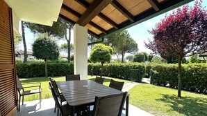 Un patio esterno con tavolo e sedie di legno, immerso in un giardino curato e delimitato da siepi.