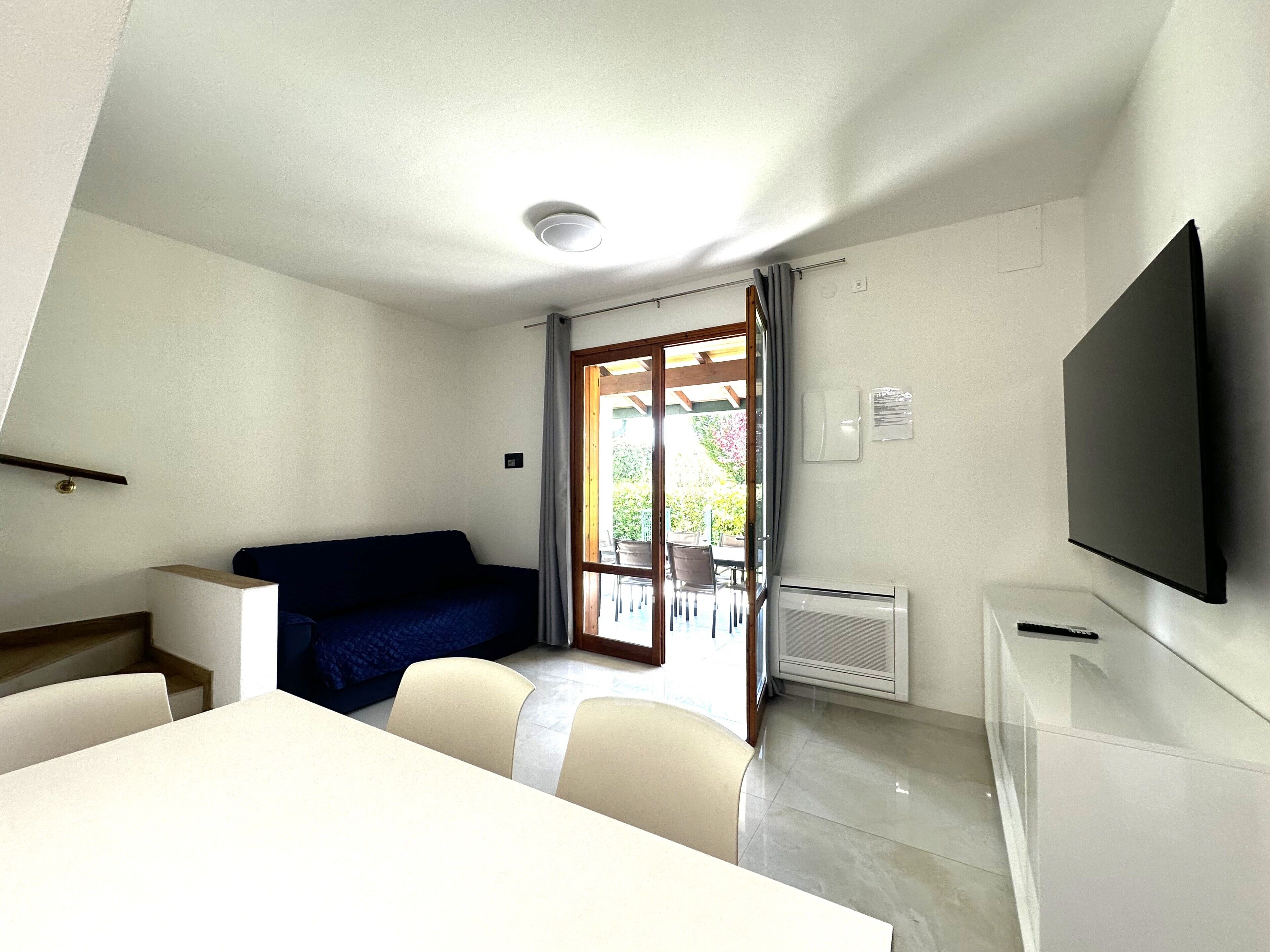 A compact room with a bed, a dining table, and chairs. There is a television mounted on the wall and a sliding glass door leading to an outdoor area.