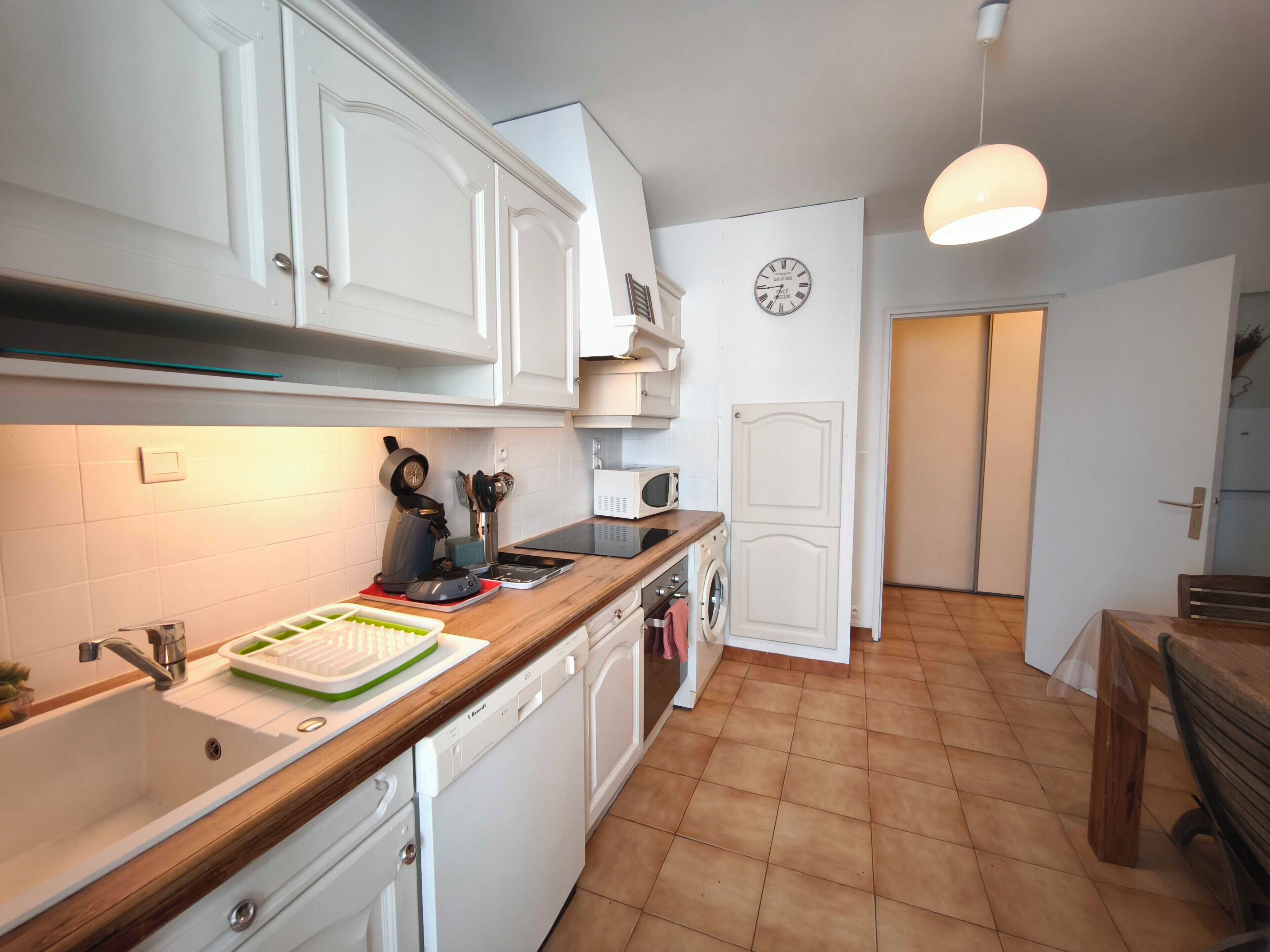 Une cuisine de taille réduite avec des meubles blancs, un plan de travail en bois, un évier, un four à micro-ondes et une horloge au mur.