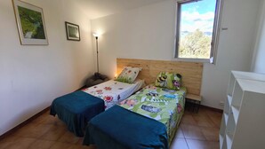 Une chambre avec deux lits, un bureau, une lampe et une fenêtre donnant sur des arbres.
