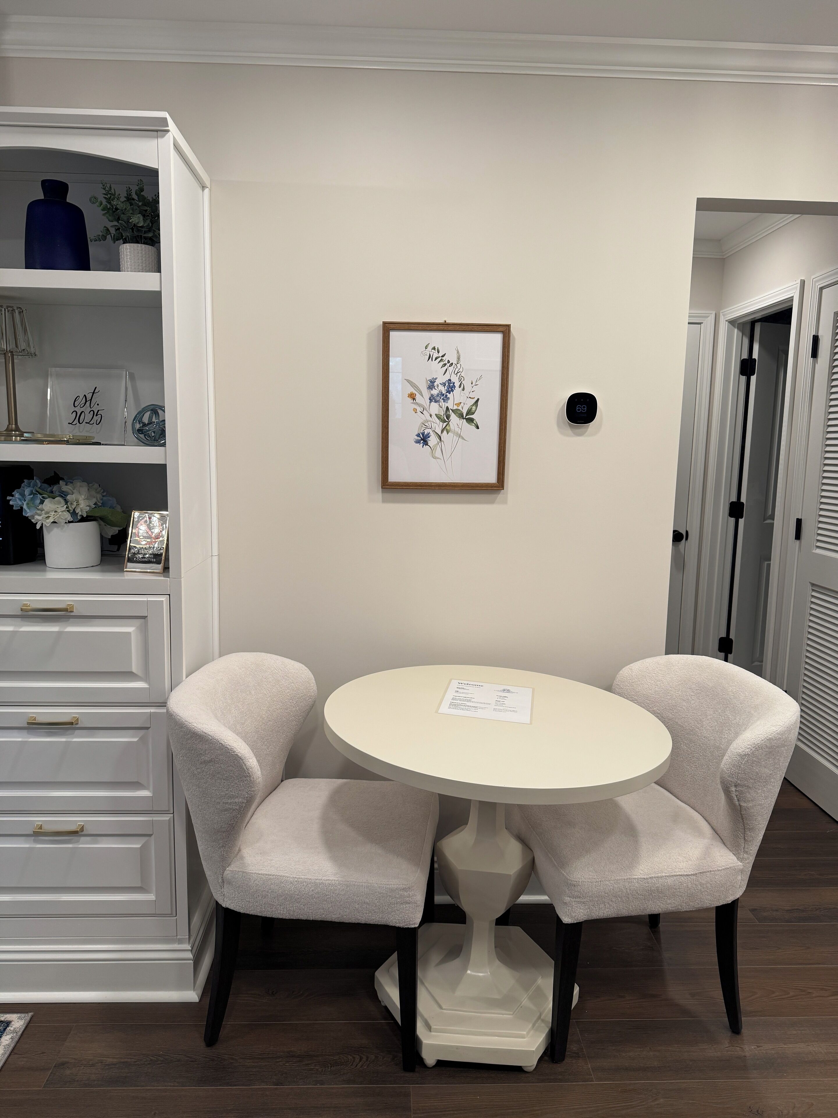 A dining area with a round table and two chairs, a white cabinet with drawers, and a framed botanical print on the wall.