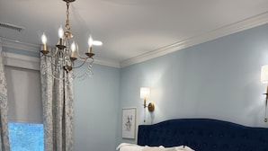 A neatly made bed with white linens and a blue headboard, a chandelier, and wall sconces.