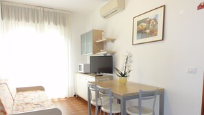 A compact living space with a kitchenette, a dining area with a table and chairs, a sofa, and a single orchid plant. - Central Bibione Apartment With Terrace (San Michele al Tagliamento)