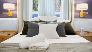 Ein ordentlich bezogenes Bett mit vielen Kissen, ein Nachttisch mit einer Lampe und ein Fenster mit Vorhängen.