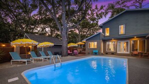 Ein Swimmingpool mit Liegestühlen und Sonnenschirmen, umgeben von einem Haus mit überdachter Terrasse.