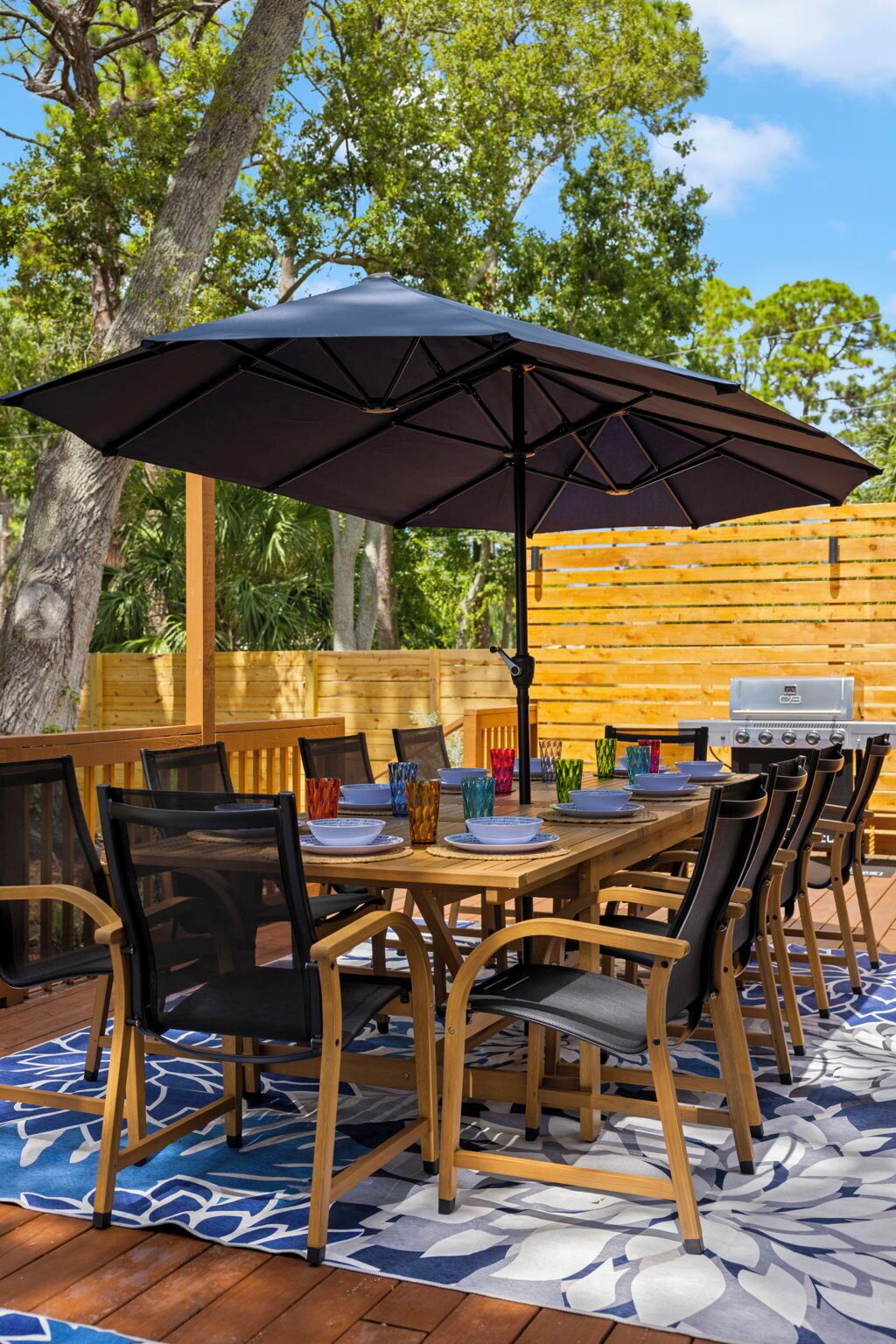 Un área de comedor al aire libre con una mesa y sillas de madera, un paraguas grande y una parrilla.