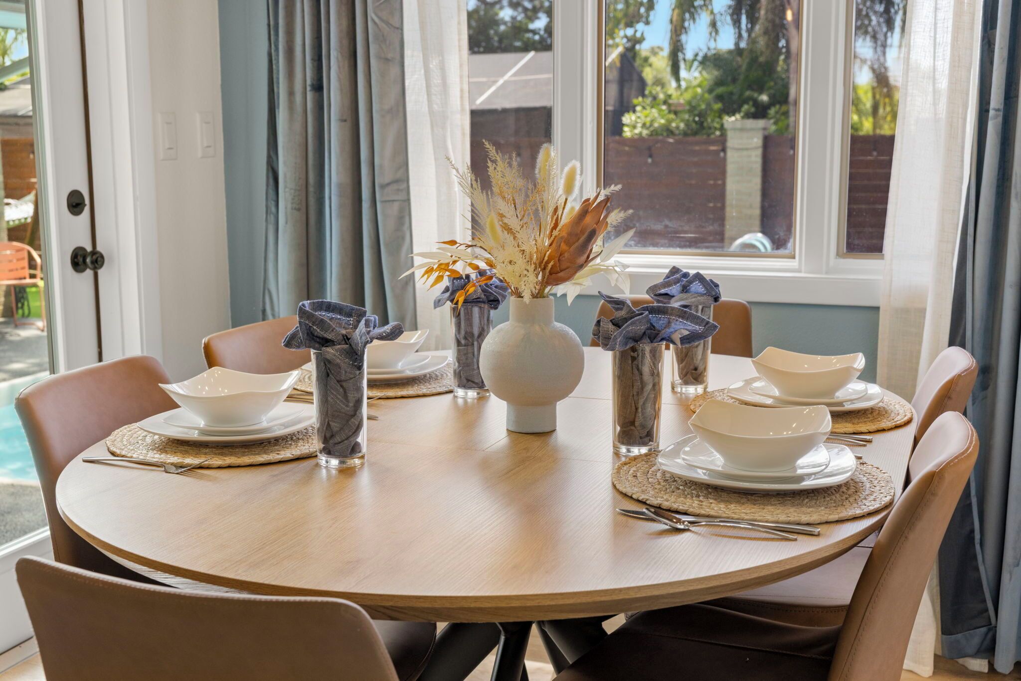 A round dining table with white plates, bowls, and a vase with dried plants.