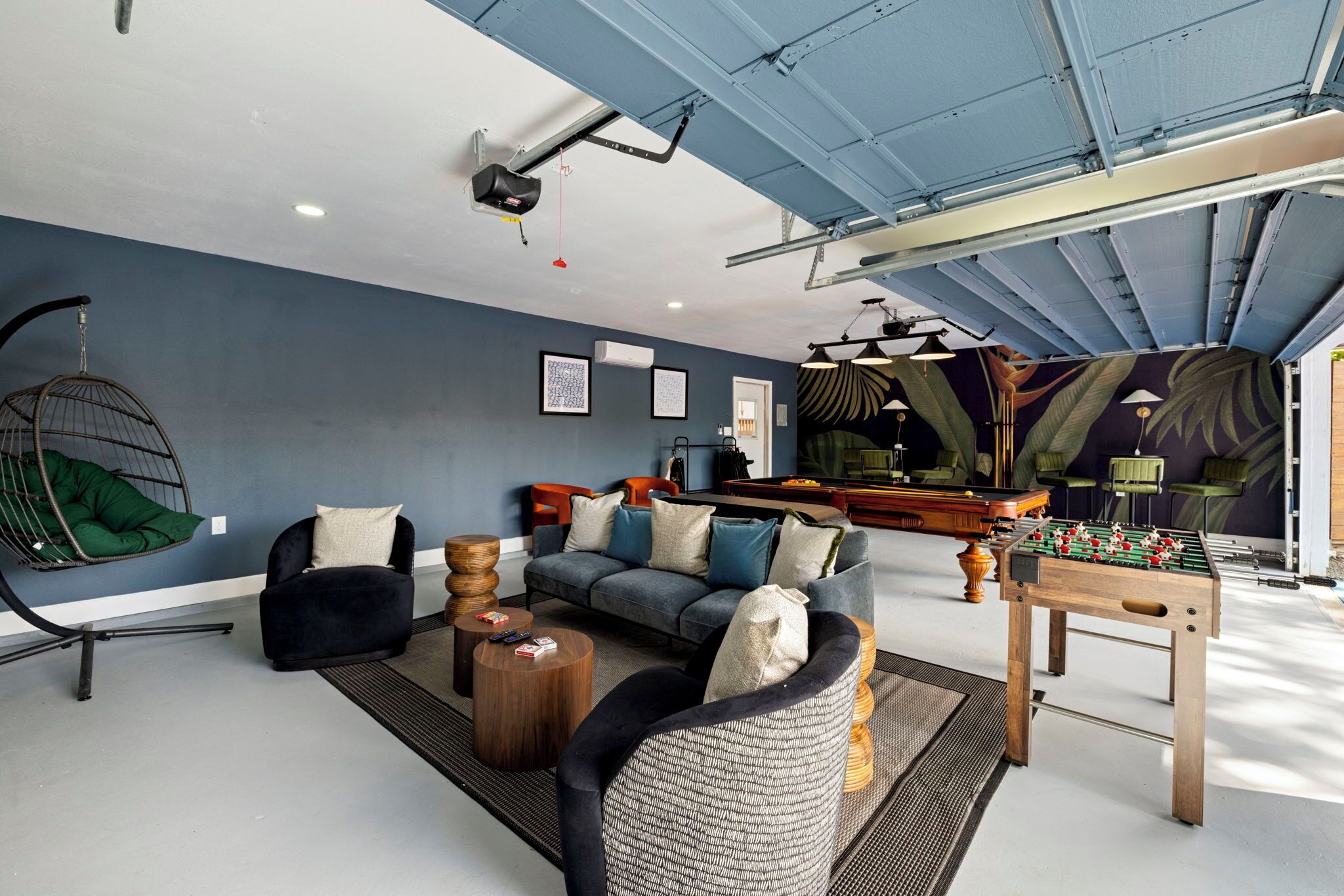 A modern living room with a sofa, armchairs, a hanging chair, a foosball table, and a ceiling fan.