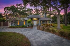 Une maison à deux étages avec une allée pavée menant à la porte d’entrée, entourée d’arbres matures et de parterres de fleurs aménagés.