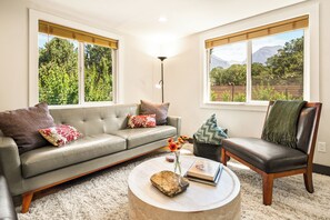 Uma sala de estar com um sofá cinzento, uma mesa de centro de madeira e uma poltrona com um cobertor verde.