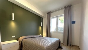 A bedroom with a bed, a bedside table, a hanging light fixture, a window with curtains, and a wall-mounted outlet. - Modern Apartment in Central Bibione (San Michele al Tagliamento)