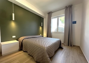 Un dormitorio con una cama, una mesita de noche, una lámpara colgante, una ventana con cortinas y un enchufe en la pared.
