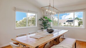 Un coin repas avec une table en bois, des chaises blanches et une grande fenêtre donnant sur des immeubles et des arbres.
