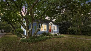 Une petite maison de plain-pied avec une porte rouge et des volets blancs, entourée d’une pelouse bien entretenue et d’arbres.