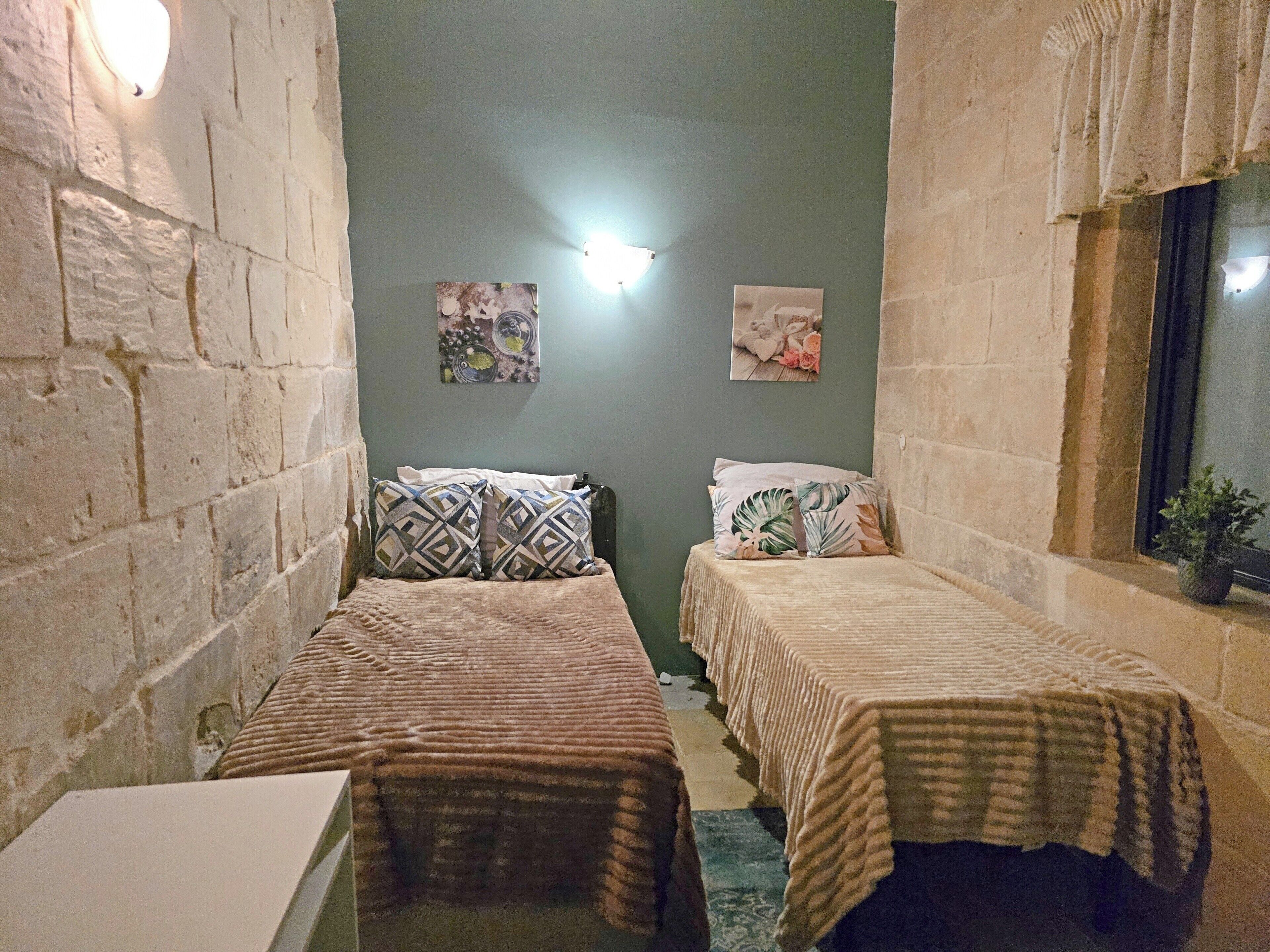 A small, dimly lit room with two single beds, a small table, and a window with curtains.