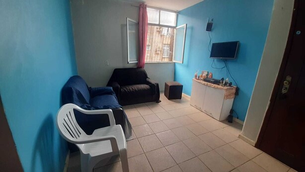 A room with blue walls, a black sofa, a white chair, a small table with items, a TV, and a window with a curtain. - Apartamento em Belém no período da COP 30 (Belem)