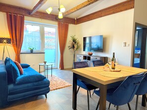A modern living room with a blue sofa, a dining table with chairs, a TV, and a small side table with a lamp. - Residence S5 - Comfort Living Apartment  (Prešov)