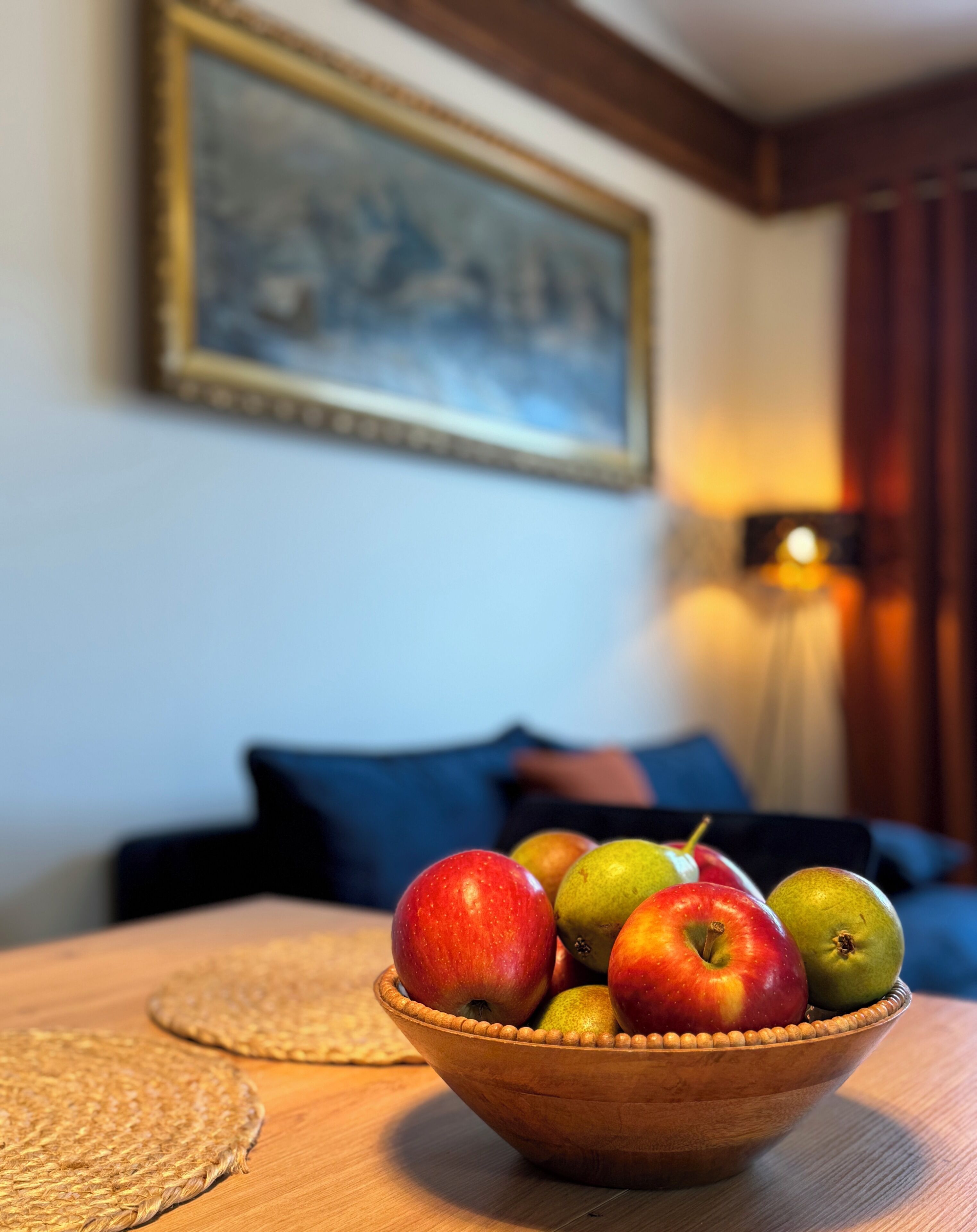 A wooden bowl with apples on a table in a room with a painting on the wall and a lamp in the background.
