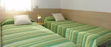 A room with two single beds, each with a green striped bedspread and white pillows. There is a small bedside table with a yellow lamp on one side.