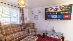 Una sala de estar con un sofá, un sillón y un televisor colocado en la pared que muestra diferentes canales.