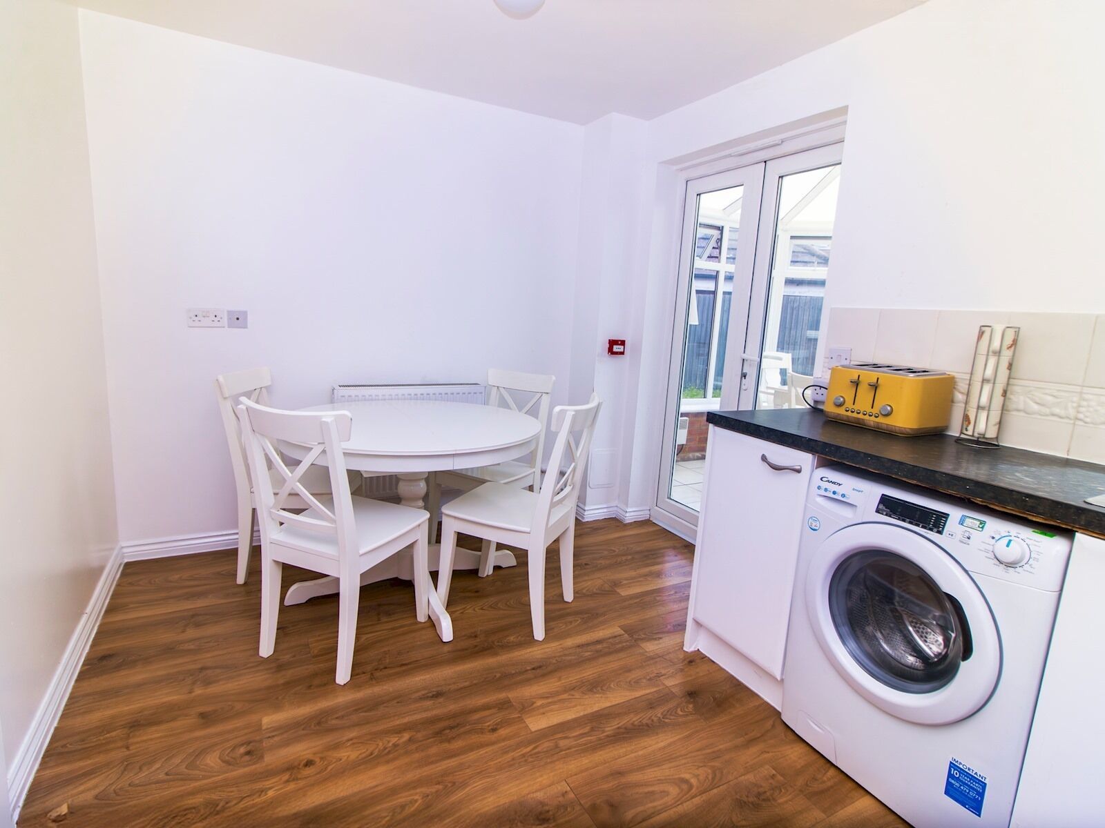 A compact kitchen with a round dining table and white chairs, a washing machine, and a window with a view of the outside.