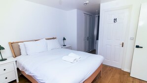 Una cama bien hecha con sábanas blancas, una mesita de noche con lámpara y un armario al fondo.
