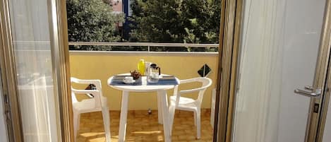 Un balcon avec une table et des chaises, une vue sur des arbres et un immeuble, et une porte donnant sur l’intérieur.