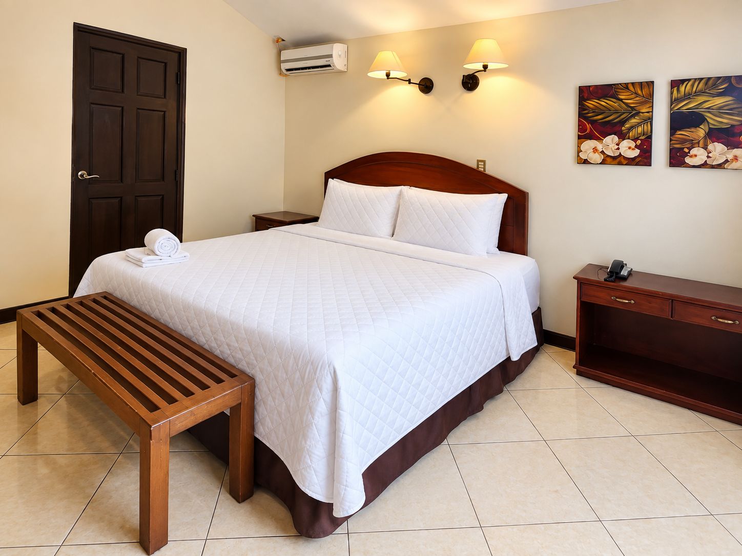 Habitación de hotel con cama, mesita de noche, banco, aire acondicionado de pared, apliques y obras de arte.