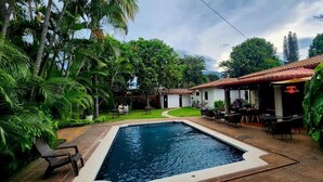 Zona de piscina con una pileta pequeña, rodeada de abundante vegetación y un patio con mobiliario exterior.