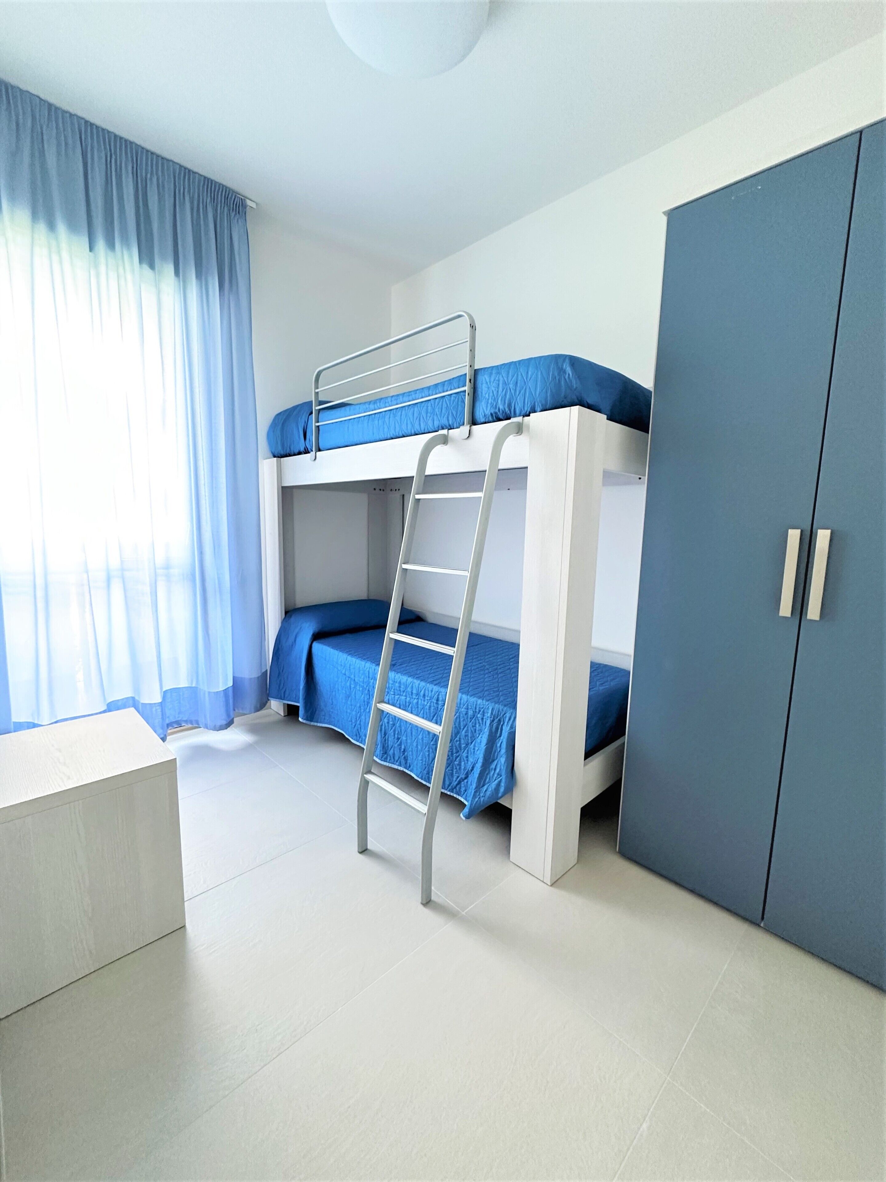 Ein Schlafraum mit Etagenbetten, einer Leiter, einem Kleiderschrank und einem Fenster mit Vorhängen.