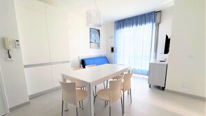 Un comedor moderno y bien iluminado con una mesa blanca y cuatro sillas, un sofá azul y una zona de cocina con un armario blanco y una lámpara colgante.