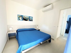 A bedroom with a bed, a nightstand, a wall-mounted air conditioner, and a painting of boats.