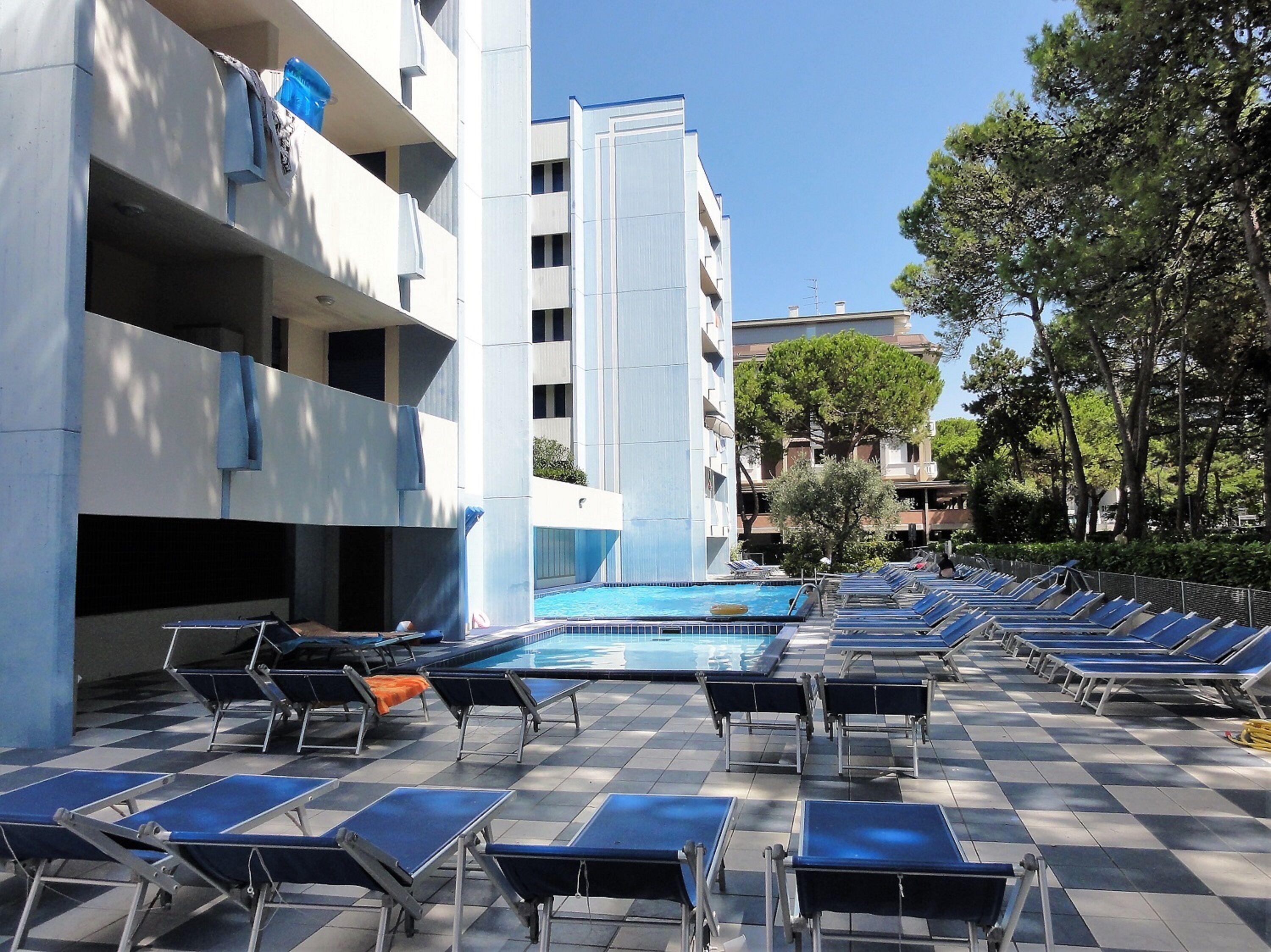 A poolside area with sun loungers and a swimming pool, surrounded by a building with balconies and trees.