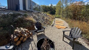 Un espacio al aire libre rústico con sillas Adirondack, un brasero y leña apilada.